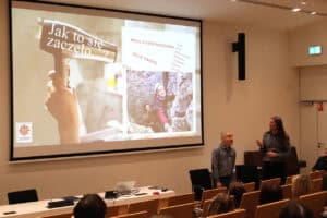 Sala wykładowa: na dużym ekranie projekcyjnym widoczny jest slajd z tytułem „Jak to się zaczęło...?", na którym widnieje duże zdjęcie dłoni trzymającej suszarkę do włosów oraz sekcje tekstowe „Moje doświadczenie, Moje pasje", ilustrowane małym zdjęciem osoby wspinającej się po skałkach. W lewym dolnym rogu jest logo Caritas. Przed ekranem stoją dwaj prelegenci: uśmiechnięty mężczyzna patrzy w lewo (Piotr Kękuś) oraz mężczyzna (Maciej Książko) z długimi włosami, gestykulujący i trzymający w dłoni przedmiot (prawdopodobnie pilot do slajdów). Widok z widowni