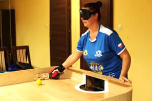 Zawodniczka w niebieskiej sportowej koszulce z biało-czerwonym akcentem i opasce na oczach, zasłaniającej widoczność, gra w showdown. Jest pochylona nad stołem, trzymając w dłoni w czerwono-czarnej rękawicy rakietkę. Przygotowuje się do uderzenia żółtej piłki, która leży przy okrągłym otworze bramkowym w drewnianym stole