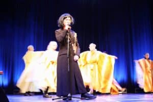 Artysta w peruce z ciemnych, krótkich włosów, ubrany w długi, czarny płaszcz stoi na środku sceny i śpiewa do mikrofonu. Za nim na tle niebieskiej kurtyny, tańczy grupa osób w luźnych, jaskrawożółtych (złotych) szatach, które są rozmyte z powodu ruchu