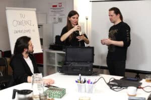 Prezentacja sprzętu: kobieta (Justyna Kazek-Góra), stojąc przy stole, unosi w rękach element sprzętu (kabel nawinięty na szpulę), pokazując go dwóm mężczyznom. Na pierwszym planie widać otwartą czarną walizkę z przenośną pętlą indukcyjną