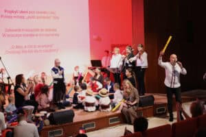 Dynamiczny moment na scenie. Od lewej: na scenie, siedzą dzieci (w tym kilkoro w tradycyjnych słomianych kapeluszach i strojach ludowych) oraz dorośli, bawią się instrumentami perkusyjnymi. Po prawej, na pierwszym planie: stoi mężczyzna w białej koszuli, śpiewa do mikrofonu i w podniesionej ręce trzyma żółtą „batutę”. Z tyłu stoi zespół: trzy kobiety, w tym dwie grają na skrzypcach. Na dużym ekranie w tle widoczny jest tekst piosenki „Przybyli ułani pod okienko".