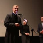 Młody ksiądz (Paweł Zazuniak) w czarnej sutannie stoi na scenie i przemawia do mikrofonu ze złożonymi dłońmi, za nim w tle stoją dwie osoby: kobieta i mężczyzna w garniturze trzymający czerwoną teczkę. Na ekranie widoczna jest część napisu „2025"