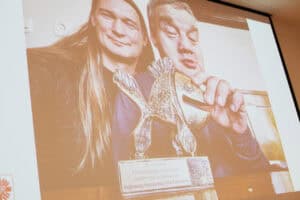 Zbliżenie na ekran projekcyjny, na którym wyświetlone jest zdjęcie dwóch mężczyzn z nagrodą filmową. Mężczyzna z lewej (Maciej Książko), z długimi włosami, lekko się uśmiecha. Mężczyzna po prawej (Piotr Kękuś) trzyma w ręku złotą statuetkę w kształcie ryby. Statuetka stoi na podstawie z plakietką, na której widnieje tekst: „20. Festiwal Filmów Optymistycznych HAPPY END w Rzeszowie / Najlepszy Niezależny Film Fabularny". Zdjęcie ma ciepły, brązowo-złoty filtr