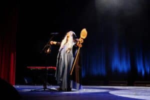 Występ Sebastiana Pacy. Artysta w kostiumie w stylu fantasy, z długimi jasnymi włosami i w błękitno-szarej, długiej szacie, stoi na scenie z szeroko rozłożonymi ramionami i głową uniesioną w górę. W jednej ręce trzyma latarnię, a w drugiej ozdobną laskę lub kostur. Tło stanowią pionowe, niebieskie kurtyny oświetlone snopami światła