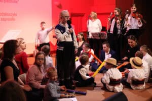 Zbliżenie na grupę dzieci, młodzieży i dorosłych na scenie oświetlonej na czerwono. W centrum uwagi stoi dziewczynka w okularach, bluzie i długiej spódnicy, mówi lub śpiewa do mikrofonu. Wokół niej siedzą inni uczestnicy, w tym dzieci w strojach ludowych i kapeluszach. W tle widoczny jest zespół: dwie skrzypaczki i kobieta w białej bluzce śpiewająca do mikrofonu