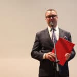 Uśmiechnięty mężczyzna (Jakub Semeniuk) w ciemnym garniturze, okularach i wzorzystym krawacie stoi na scenie i przemawia do mikrofonu, trzyma w lewej ręce jaskrawoczerwoną teczkę