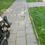 Ulica Weteranów w Lublinie. Wąski, szary chodnik z betonowych płyt biegnie w głąb kadru w perspektywie zbieżnej. Po prawej stronie odchodzą od niego wąskie boczne ścieżki, których dalszy przebieg nie jest widoczny. W lewym dolnym rogu kadru widoczna jest prawa strona elektrycznego wózka inwalidzkiego z kołami. Po obu stronach chodnika znajdują się trawniki porośnięte zieloną trawą. Trawnik po lewej porasta szpaler drzew, za którymi znajduje się jezdnia i rząd samochodów.