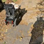 Ulica Józefa Sowińskiego w Lublinie. Nasłoneczniona nawierzchnia chodnika ukazuje cień osoby stojącej po prawej stronie. Chodnik z licznymi ubytkami i nierównościami pokrywają suche, żółtobrązowe liście, znajduje się na nim widoczny tyłem elektryczny wózek inwalidzki, w którym siedzi kobieta z długimi włosami.