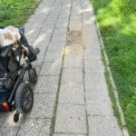 Ulica Weteranów w Lublinie. Wąski, szary chodnik z betonowych płyt biegnie w głąb kadru w perspektywie zbieżnej. Po prawej odchodzi jedna boczna ścieżka, której dalszy przebieg pozostaje niewidoczny. Po lewej stronie kadru widać prawą część elektrycznego wózka inwalidzkiego z kołami, zbliżającego się do ubytku w chodniku – dziury wypełnionej brudnym piaskiem. Po obu stronach chodnika rozciągają się trawniki porośnięte zieloną trawą.