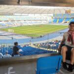 Po lewej stronie kadru na zadaszonym sektorze stadionu znajduje się młody mężczyzna na wózku inwalidzkim. Ubrany jest w czapkę z daszkiem, szalik kibica Polski, krótkie spodenki i sportową koszulkę bez rękawów. Za nim widać murawę stadionu i sektory wokół, a na niższym poziomie, trybuny częściowo wypełnione ludźmi