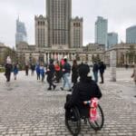 Plac przed Pałacem Kultury i Nauki w Warszawie. Na pierwszym planie tył wózka inwalidzkiego, w którym siedzi mężczyzna ubrany na czarno; na oparciu wózka przewiązany biało-czerwony szalik kibica. W tle po chodniku spacerują grupy osób w cieplejszej odzieży, wiele z nich ma elementy patriotyczne, w tym dwaj mężczyźni z biało-czerwonymi opaskami na lewych przedramionach oraz osoby z powiewającą biało-czerwoną flagą Polski. Zdjęcie wykonano przy pochmurnej aurze, drzewa częściowo bez liści