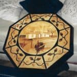 Ośmiokątny blat drewnianego stołu zdobionego techniką intarsji. Po środku motyw domu w jasnych kolorach; dookoła ornament roślinny