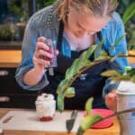 Młoda kobieta w dżinsowej koszuli i czarnym fartuchu przyprawia pucharek deserowy wypełniony bitą śmietaną. Spogląda uważnie w dół, trzymając młynek nad pucharkiem. Deser leży na białej desce do krojenia na drewnianym blacie, a obok niego leży nóż.