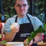 Uśmiechnięty mężczyzna w okularach i czarnym fartuchu z białym napisem „SZWEDZKI STÓŁ” trzyma w prawej ręce ciasto, a drugą ręką podtrzymuje dużą białą miskę.