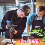 Dwóch kucharzy w fartuchach. Jeden z nich używa rękawa cukierniczego i tworzy małe, okrągłe desery, podczas gdy drugi obserwuje jego pracę.