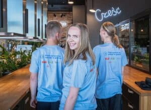 Trzy osoby w identycznych, jasnoniebieskich koszulkach z napisem „Mam apetyt na sukces w gastro!”. Kobieta w środku stoi twarzą do kamery i uśmiecha się. Po obu jej stronach znajdują się dwie osoby z głowami zwróconymi tyłem. Za nimi widoczny jest neonowy napis „Cześć”.