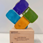 Statua-nagroda składająca się z czterech prostokątów ceramicznych w kolorach niebieskim, fioletowym, zielonym i żółtym, ułożonych w kwadrat i osadzonych na podstawie z jasnego drewna z napisem: NAGRODA KARTY RÓŻNORODNOŚCI 2025. KATEGORIA: DEI W BIZNESIE. PODKATEGORIA PARTNERSTWO. Lublin Skills Up! Apetyt na sukces w gastro Ingka Centers Polska sp. z o.o. KARTA RÓŻNORODNOŚCI KOORDYNOWANA PRZEZ FORUM ODPOWIEDZIALNEGO BIZNESU.