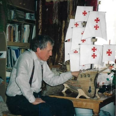 W pokoju przy stole ustawionym obok okna, siedzi starszy mężczyzna w białej koszuli i granatowych spodniach na szelkach. Jedną ręką podpiera kolano, drugą dotyka stojącego przed nim modelu żaglowca. W tle widać regał pełen książek