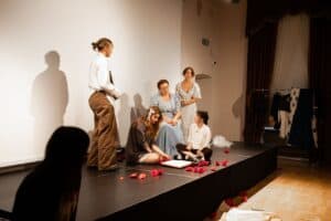 Pięć młodych kobiet w strojach scenicznych pozuje na scenie. Część z nich stoi, część siedzi; na białej ścianie w tle widoczne są cienie ich sylwetek