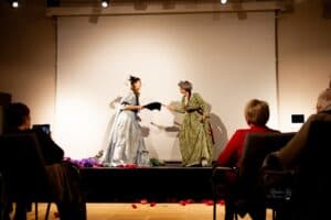 Widok sceny z perspektywy widowni: dwie kobiety w długich sukniach stylizowanych na stroje z epoki, stoją zwrócone do siebie przodem. W wyciągniętych dłoniach trzymają wachlarze, a ich gesty i mimika sugerują napiętą rozmowę; w tle na białej ścianie widoczne są cienie ich sylwetek