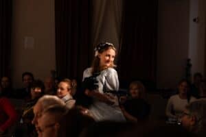 Wśród siedzącej publiczności stoi młoda kobieta w błękitnej sukni stylizowanej na strój z epoki. Oświetlona reflektorem odwraca głowę za siebie, podczas gdy reszta widowni pozostaje w cieniu