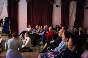 Publiczność siedzi na krzesłach ustawionych w półokręgu, zwrócona w stronę sceny znajdującej się poza kadrem. Na drewnianej podłodze przed rzędami siedzeń siedzą dwie młode kobiety, z których jedna oświetla latarką przestrzeń przed sobą; w tle widoczne są trzy okna zasłonięte bordowymi aksamitnymi kotarami