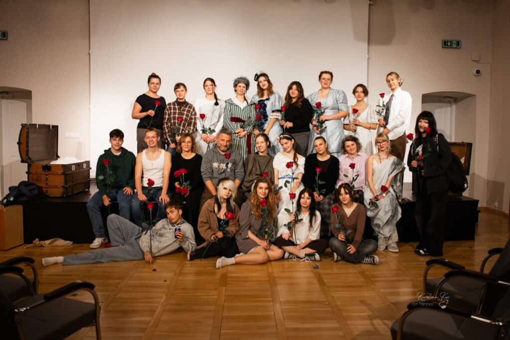Pamiątkowe zdjęcie grupowe uczestników projektu „Tableau vivant” – młodzieży z Zespołu Szkół nr 2 w Lubartowie. Dwudziestoczteroosobowa grupa pozuje na scenie w trzech rzędach, część w kostiumach scenicznych, część we współczesnych ubraniach, każdy z czerwoną różą w dłoni