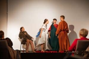 Na scenie po lewej stronie siedzi bosa kobieta w brązowej tunice z obfitym, pomarszczonym materiałem i odsłoniętymi nogami. Obok niej rozgrywa się scena między młodą kobietą i dwoma młodzieńcami, ubranymi w stroje przypominające epokę starożytności; wszyscy są boso. Kobieta pochyla się w stronę mężczyzn z otwartymi ustami, jakby mówiła z pretensją lub krzyczała