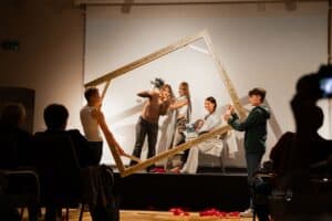 Widok sceny z perspektywy widowni. Trzy kobiety tworzą żywy obraz: dwie ubrane współcześnie, jedna z nich pokazuje gest „okej”, druga trzyma bukiet kwiatów i ma na ramieniu narzuconą tunikę; trzecia, w tunice, siedzi nieruchomo z kwiatami na kolanach i lekko się uśmiecha. Po obu stronach sceny dwaj młodzieńcy trzymają ozdobną ramę obejmującą całą grupę