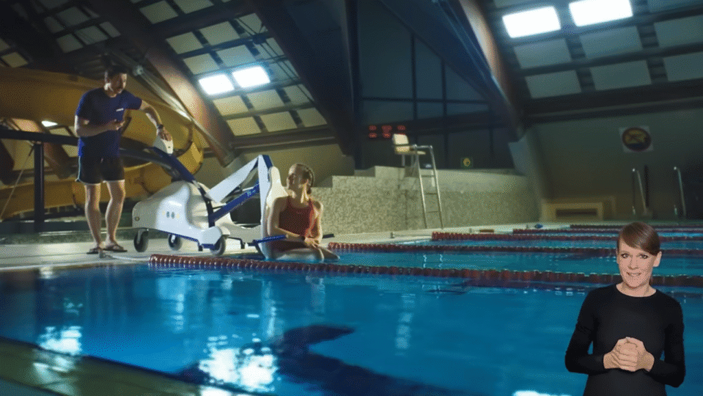 Kobieta siedzi przy krytym basenie, korzystając z podnośnika basenowego, w pobliżu stoi mężczyzna, a w prawym dolnym rogu widać tłumacza języka migowego.