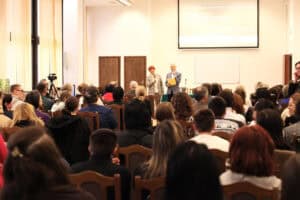 Panoramiczne ujęcie wnętrza sali wykładowej. Kobieta w kraciastym żakiecie (Maria Pietrusza-Budzyńska) i mężczyzna w garniturze (prof. Wojciech Otrębski) stoją trzymając mikrofony i przemawiają do licznie zgromadzonej publiczności. Na ścianie za nimi widać duży ekran projekcyjny z napisem "KONFERENCJA NAUKOWO-SZKOLENIOWA", a pod nim białą tablicę. Uczestnicy, widoczni z tyłu, siedzą na drewnianych krzesłach