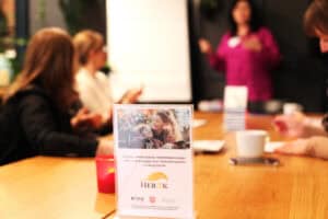 Na drewnianym stole konferencyjnym stoi biała tabliczka informacyjna w przezroczystej ramce ze zdjęciem uśmiechniętej kobiety przytulającej dziecko na wózku inwalidzkim i tekstem: „Projekt Matka dziecka niepełnosprawnego – siła i moc każdego dnia realizowany przez Fundację HERPIK”. U dołu tabliczki logotypy partnerów: ROPS Lublin, Województwo Lubelskie oraz PFRON. W rozmytym tle widać uczestniczki spotkania siedzące przy stole oraz prowadzącą w bluzce koloru fuksji