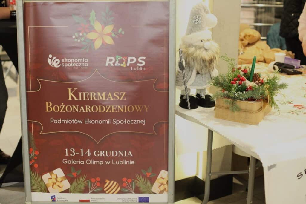 Plakat informacyjny na stojaku o treści: „Kiermasz Bożonarodzeniowy Podmiotów Ekonomii Społecznej, 13-14 grudnia, Galeria Olimp w Lublinie”. Na plakacie widoczne logotypy: Ekonomia Społeczna, ROPS Lublin oraz barwy unijne. Obok plakatu, na stole nakrytym białym obrusem, stoi dekoracyjna figurka skrzata w szarym ubraniu oraz stroik bożonarodzeniowy z igliwia z zieloną świecą.