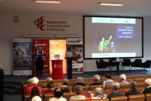 Szerokie ujęcie auli wykładowej podczas konferencji. Na środku sceny stoi uśmiechnięta kobieta (dr Marta Komorska) z teczką w ręku. Po jej lewej stronie widoczne są kolorowe banery reklamowe, a po prawej duży ekran projekcyjny z napisem: „Konferencja Senioralni Liderzy Dostępności, Lublin, 16 grudnia 2025 r.”. W dolnej części zdjęcia widoczne są rzędy krzeseł zajęte przez słuchaczy, głównie osoby starsze, widziane od tyłu. Na ścianie nad sceną widnieje napis: „Wyższa Szkoła Przedsiębiorczości i Administracji”.