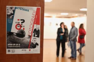 Plakat informacyjny o wystawie fotograficznej "Czego nie rozumiesz? Jestem taki jak Ty" w Janowskim Ośrodku Kultury, wiszący na drewnianych drzwiach, z częściowo widoczną, rozmytą salą wystawową w tle, gdzie widać sylwetki kilku osób