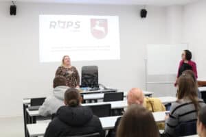 Wnętrze sali wykładowej. Kobieta (Ewelina Orzechowska) w ciemnej, wzorzystej bluzce stoi za biurkiem i przemawia do grupy młodych ludzi siedzących w rzędach białych ławek. Za nią na tle jasnej ściany widoczne jest logo "ROPS Lublin" oraz herb Województwa Lubelskiego. Obok ekranu stoi tablica suchościeralna