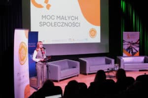 Od lewej: baner stojący Aktywne Małe Społeczności, utrzymany w kolorach białym i pomarańczowym. Przed banerem kobieta (Małgorzata Kotowska), w biznesowym stroju z stoi na scenie przy statywie z mikrofonem i przemawia. W tle widoczny jest duży ekran z hasłem "Moc Małych Społeczności" oraz trzy szare sofy. Na pierwszym planie widoczne są tyły głów publiczności, która słucha prelekcji w zaciemnionej sali
