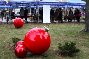 Na pierwszym planie od lewej, na trawie leżą cztery błyszczące, gigantyczne bombki świąteczne w kolorze czerwonym. Trzy z nich są mniejsze, a jedna, największa, ma widoczny złoty uchwyt. W tle widoczny jest długi szereg niebieskich namiotów z białym napisem „Uniwersytet Marii Curie-Skłodowskiej w Lublinie", pod którymi stoją ludzie przeglądający stoiska kiermaszu plenerowego