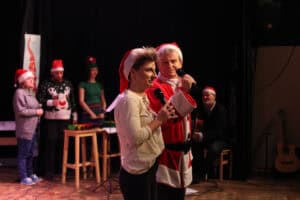 Na słabo oświetlonej scenie, dwoje prowadzących (Karolina Czajka, Michał Stanowski) w świątecznych czapkach (kobieta w beżowej bluzce, mężczyzna w kostiumie Mikołaja) stoi obok siebie i przemawia do mikrofonów. Kobieta patrzy na wprost, a mężczyzna w prawo. W tle, stoją trzy inne osoby: jedna w szarej bluzie, druga w swetrze z reniferem, trzecia w zielonej koszulce. Na prawo widoczny jest mężczyzna z gitarą i gryf gitary akustycznej opartej o ścianę.