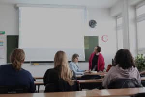 Sala wykładowa. Trzy osoby siedzą przy ławkach, tyłem do obiektywu, naprzeciwko dwóch kobiet, z których jedna siedzi, a druga stoi przy biurku. Prowadząc Anna Bieganowska-Skóra w różowym kardiganie patrzy na duży ekran, który znajduje się na ścianie z tyłu. Przy biurku siedzi Dorota Krać ubrana w niebieski żakiet i pochyla się nad laptopem. Salę wypełnia jasne światło zastane, z okien po prawej stronie kadru