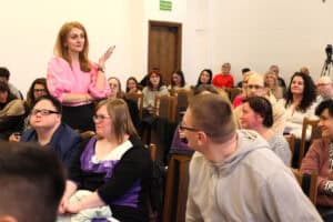 Kobieta o długich, blond włosach (dr Monika Zima-Parjaszewska), ubrana w różową bluzkę, stoi po lewej stronie kadru z uniesioną dłonią i gestykuluje w stronę słuchaczy. W rzędach przed nią siedzi kilkanaście osób, w tym na pierwszym planie mężczyzna w szarej bluzie, odwrócony w stronę kobiety oraz inna kobieta w fioletowo-czarnym ubraniu. Z tyłu sali widać ciemne, drewniane drzwi i fragment kamery na statywie