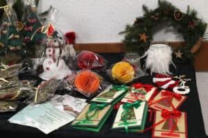 Stół z dekoracjami bożonarodzeniowymi, na którym widać ręcznie robione kartki, bałwanka w folii, choinki z materiału oraz wieniec z igliwia z suszonymi pomarańczami.