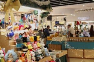 Schodkowa ekspozycja z ręcznie robionymi zabawkami na szydełku. Na stopniach ustawiono dziesiątki kolorowych maskotek: lalki w zimowych strojach oraz zwierzątka, m.in. misie, króliki i postacie przypominające Kubusia Puchatka. Nad stoiskiem wiszą kolorowe breloki i ozdoby w kształcie gwiazdek. W tle widać pasaż galerii handlowej z innymi stoiskami kiermaszowymi, przy których stoją wystawcy i klienci.