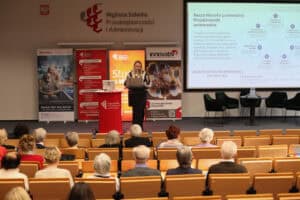 Wystąpienie prelegentki – kobiety w okularach i pasiastej koszuli (Karolina Kowalczyk). Na ekranie za nią widoczny jest slajd o tytule „Nasza filozofia przewodnia: Projektowanie uniwersalne”. Slajd zawiera schemat kołowy opisujący „7 zasad projektowania uniwersalnego”, m.in. elastyczność w użytkowaniu, prostą i intuicyjną obsługę oraz niski poziom wysiłku fizycznego.