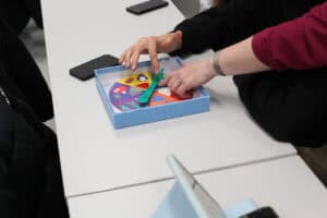 Zbliżenie na grę planszową w niebieskim pudełku, leżącą na jasnym stole. Dwie pary rąk – jedna w czarnej bluzie, druga z ciemnym rękawem – wspólnie obsługują zieloną wskazówkę na kolorowym kole. W tle widoczne są dwa smartfony