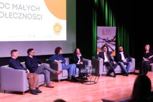 Sześć osób (m.in.: Piotr Stec, Gabriela Figura, Aleksander Jurzysta, Ewa Kalbarczyk, Piotr Marzęda) siedzi na szarych kanapach podczas panelu dyskusyjnego. Osoby są pogrążone w rozmowie, wymieniają się spojrzeniami na scenie oświetlonej ciepłym światłem. Jedna z kobiet trzyma mikrofon i przemawia. Za nimi, po lewej stronie, widoczny jest ekran z fragmentem hasła „Moc Małych Społeczności", a po prawej baner informacyjny „Moc Małych Społeczności”. Na pierwszym planie widać niewielki fragment publiczności