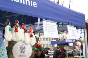 Fragment niebieskiego namiotu. Pod daszkiem wisi pięć ręcznie robionych ozdób z białej wełnianej włóczki, imitujących postacie z brodami, czerwonymi nosami i kolorowymi czapkami. Obok wiszą kartki informujące: „Ogród Botaniczny UMCS prowadzi zbiórkę na rzecz Dziennego Ośrodka Adaptacyjnego dla Dzieci i Młodzieży z Niepełnosprawnością Intelektualną przy ulicy Poturzyńskiej 1 w Lublinie”.