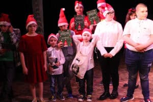 Grupa kilkorga dzieci i nastolatków w świątecznych czapkach stoi na scenie. W centralnej części dwóch starszych chłopców trzyma dyplomy z zielonym nadrukiem i napisem „DYPLOM". Najmniejszy chłopiec pośrodku stoi z szeroko uniesionymi rękami, ma zawieszoną na szyi beżową torbę ze wzorem. Po prawej stronie, obok niego, stoi wyższy chłopiec w okularach, białej koszuli i czarnych spodniach, ze złożonymi rękami. Po lewej stronie widać dziewczynkę w czerwonej, aksamitnej sukience. Wszyscy są oświetleni ciepłym światłem.