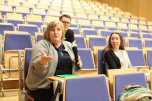 Zbliżenie na uczestników siedzących w auli wykładowej z niebieskimi fotelami i drewnianymi pulpitami. Na pierwszym planie kobieta w szarym kardiganie trzyma czarny mikrofon i wykonuje gest dłonią, przemawia do zgromadzonych. Obok niej siedzi inna kobieta w czarno-szarej bluzce, a w tle, między nimi, widoczny jest mężczyzna w okularach. Tło zdjęcia stanowią puste rzędy foteli wznoszące się ku górze auli