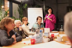 Grupa uczestniczek przy drewnianym stole. Prowadząca w fuksjowej koszuli stoi w tle obok białego flipcharta z napisami „moje talenty”, „moje wartości”, „moje osiągnięcia”. Na stole butelki wody, kubki, świeczniki i pudełka z chusteczkami. Po lewej fragment przeszklonej ściany z roślinami.